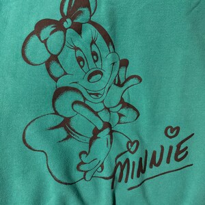 May include: Teal-colored sweatshirt featuring a brown illustration of Minnie Mouse. The design includes Minnie's face, bow, and the word "Minnie" in a cursive font with a heart above the "i".