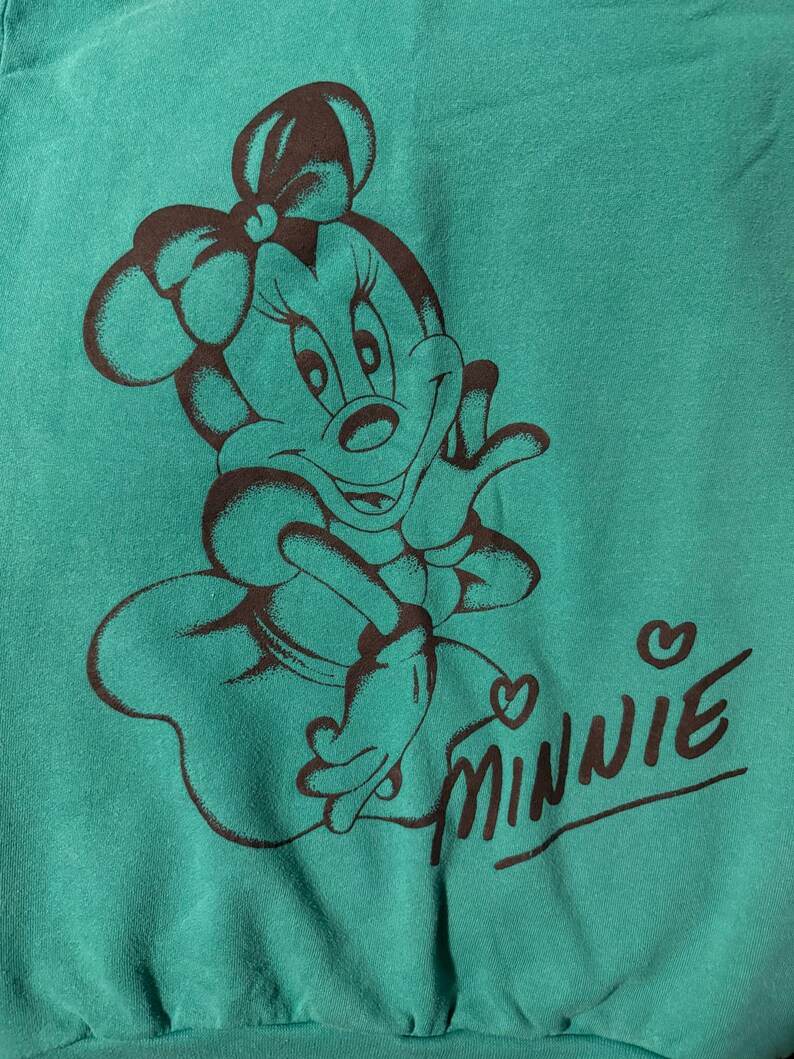 May include: Teal-colored sweatshirt featuring a brown illustration of Minnie Mouse. The design includes Minnie's face, bow, and the word "Minnie" in a cursive font with a heart above the "i".