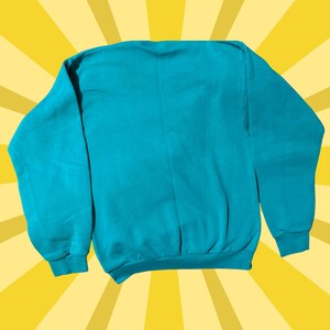 May include: A teal-colored sweatshirt with a crew neck and long sleeves. The sweatshirt is made of a soft material and has ribbed cuffs and hem. The background is a yellow burst pattern.