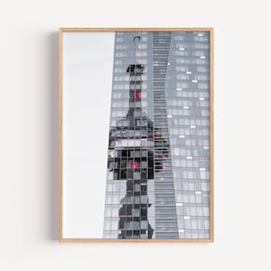 May include: A framed print featuring a modern architectural photograph. The image showcases a tall, glass-clad skyscraper with a mirrored reflection of a structure with red accents. The print is encased in a light-colored wooden frame.