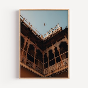 May include: A framed print showing an architectural structure with detailed features. The image captures a low-angle view of a building with arched windows and decorative balconies. A bird flies in the blue sky above the building. The building is a warm, earthy tone.
