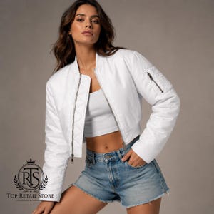 May include: A white cropped bomber jacket featuring a quilted texture, a front silver zipper closure, and a zippered utility pocket on the left sleeve. The jacket is worn over a white crop top and blue denim shorts with a raw hem finish.