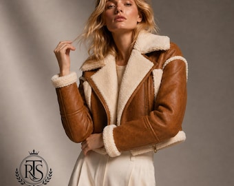 May include: A cropped tan jacket with a wide cream-colored faux shearling collar, cuffs, and hem trim. The jacket features a smooth tan exterior, long sleeves, and a front zipper closure, paired with plain cream-colored trousers.