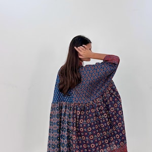 May include: A patterned dress in shades of blue, red, and white. The dress features a mix of geometric and floral designs. The sleeves and hem have a red border. The dress is loose-fitting and appears to be made of a lightweight fabric.