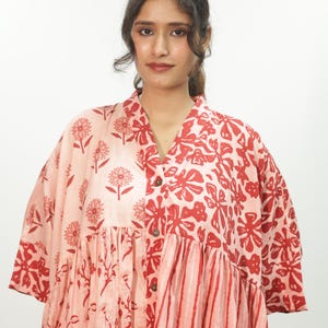 Peut inclure: Une chemise boutonnée ample présentant un motif patchwork de fleurs, de végétaux et de rayures dans des tons rose clair et rouge vif. Le vêtement possède un col en V, des manches courtes larges et une taille froncée créant une silhouette volumineuse.