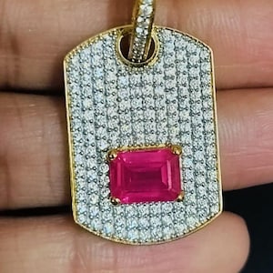 May include: Gold-tone pendant featuring a rectangular, bright pink gemstone. The pendant is covered in small, clear stones and has a gold bail. The design is a dog tag shape, with a gold border.