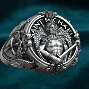 Puede incluir: Un anillo de plata detallado con San Miguel con alas y una espada, rodeado por las palabras "SAINT MICHAEL". El anillo tiene un diseño ornamentado con una cruz en el lateral, mostrando intrincados trabajos en metal y una detallada representación del santo.