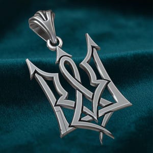 May include: A silver pendant featuring the Tryzub, the national emblem of Ukraine. The pendant has a detailed, three-pronged trident design with a loop for a chain. The metal has a polished finish, and the background is a teal color.