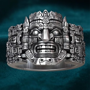Op de afbeelding: Een zilverkleurige ring met een gedetailleerd Azteeks maskerontwerp. De ring toont een centraal gezicht met een open mond, geflankeerd door ingewikkelde gravures en een kroon. Het ontwerp is zeer gedetailleerd en driedimensionaal.