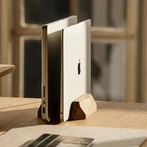 Op de afbeelding: Een zilveren laptop wordt rechtop gehouden in een houten standaard. De standaard is gemaakt van lichtgekleurd hout met een geometrisch ontwerp. De laptop heeft een zwart Apple-logo op de achterkant. De standaard staat op een houten bureau.