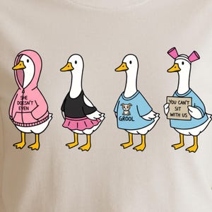 May include: A cream-colored t-shirt featuring four cartoon ducks, each dressed in different outfits. One duck wears a pink hoodie with the text "She Doesn't Even." Another wears a black top and pink skirt. A third duck has a blue shirt with a dog graphic and the word "GROOL." The last duck holds a sign that says "You Can't Sit With Us."