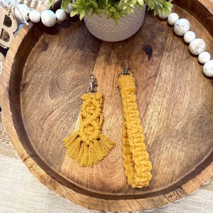 May include: Mustard yellow macrame keychain and wristlet with silver-tone hardware. The keychain has a tassel design, while the wristlet is a looped design. Both are displayed on a wooden tray with decorative elements.