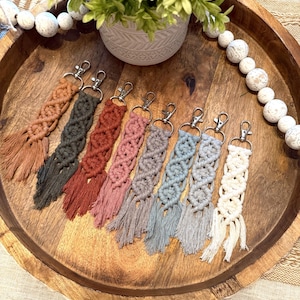 May include: A collection of handmade macrame keychains in various colors, including brown, green, red, pink, gray, blue, and cream. Each keychain features a woven design and a tassel, attached to a silver-tone clasp. The keychains are displayed on a wooden tray.