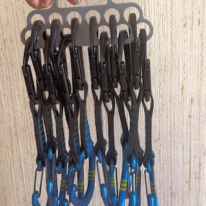 Könnte beinhalten: Eine Sammlung von Kletter-Expressschlingen. Jede Expressschlinge hat einen schwarzen Karabiner, der mit einem blauen Gurtband mit gelben und grünen Akzenten verbunden ist. Die Expressschlingen sind an einem silbernen Metallbügel befestigt.