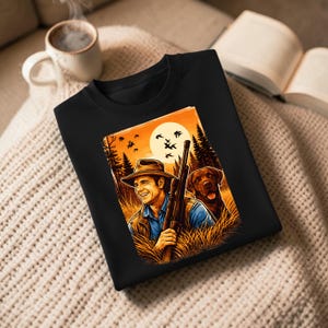 Pode incluir: Camisola preta com uma ilustração colorida de um homem com um rifle e um cão num campo. A obra de arte retrata um pôr do sol com pássaros a voar, árvores e erva alta. O homem está a usar um chapéu e a sorrir.