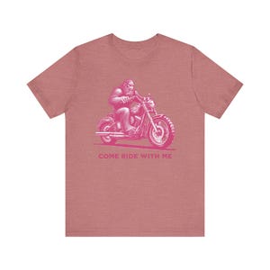 May include: A dusty rose t-shirt featuring a pink illustration of a Bigfoot riding a motorcycle. The text "COME RIDE WITH ME" is printed below the motorcycle. The design is a playful take on biker culture.