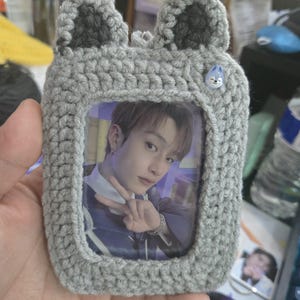 Puede incluir: Un marco de fotos de ganchillo gris con una foto de una persona en su interior. El marco tiene dos orejas grises en la parte superior y un pequeño botón azul con una cara. El marco es rectangular y parece hecho a mano.
