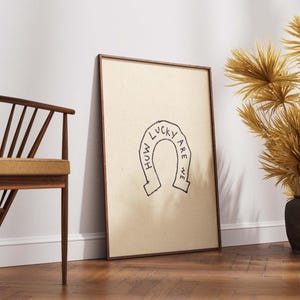 May include: A framed print with a tan background and a black horseshoe design with the words "HOW LUCKY ARE WE". The frame is brown. A wooden chair and decorative dried plants in dark vases are also visible.