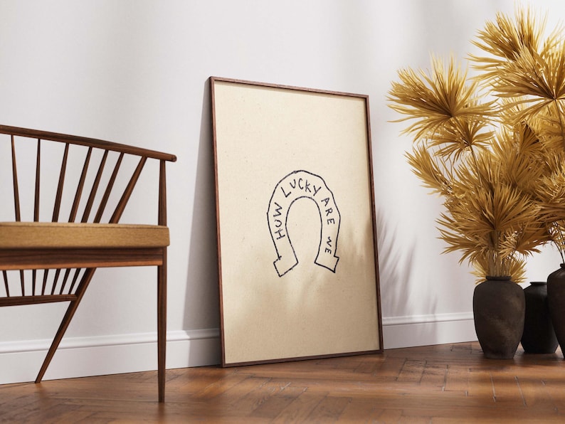 May include: A framed print with a tan background and a black horseshoe design with the words "HOW LUCKY ARE WE". The frame is brown. A wooden chair and decorative dried plants in dark vases are also visible.