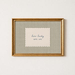May include: A rectangular gold frame containing a beige and white gingham patterned background. Centered within the frame is a cream-colored rectangular card with scalloped edges featuring the dark blue text how lucky are we.