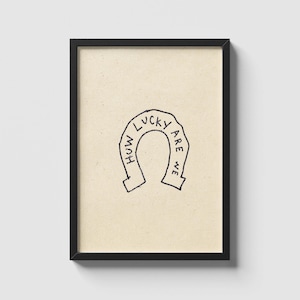 May include: A framed art print featuring a horseshoe illustration with the words "How Lucky Are We" in a handwritten style. The artwork is in black on a beige background, encased in a black frame.