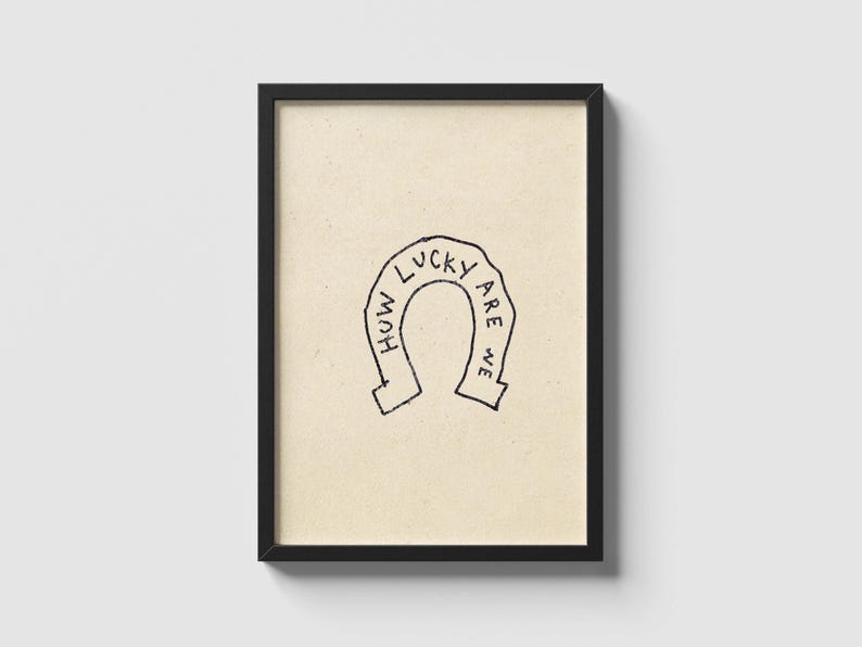 May include: A framed art print featuring a horseshoe illustration with the words "How Lucky Are We" in a handwritten style. The artwork is in black on a beige background, encased in a black frame.