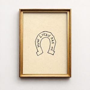 May include: A framed print with a gold-colored frame. The artwork features a black horseshoe illustration with the words "How lucky are we" written inside the horseshoe. The background is a light tan color.