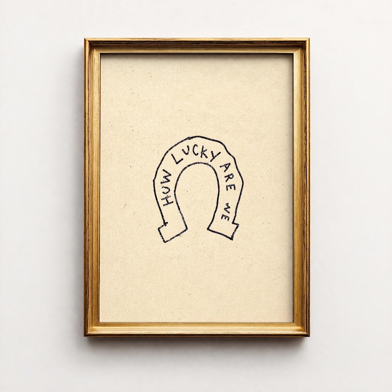 May include: A framed print with a gold-colored frame. The artwork features a black horseshoe illustration with the words "How lucky are we" written inside the horseshoe. The background is a light tan color.