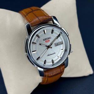 May include: A silver Seiko 5 automatic watch with a brown leather strap. The watch face is silver with black hands and markers, and the day and date are displayed at the 3 o'clock position. The watch is displayed on a white pillow.