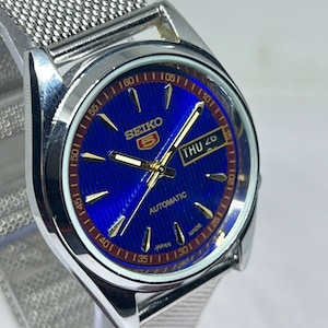 Könnte beinhalten: Eine silberfarbene Seiko Automatik-Armbanduhr mit blauem Zifferblatt und Mesh-Armband. Das Zifferblatt hat goldfarbene Zeiger und Markierungen, mit Anzeige von Tag und Datum. Die Uhr ist mit "Made in Japan" gekennzeichnet.