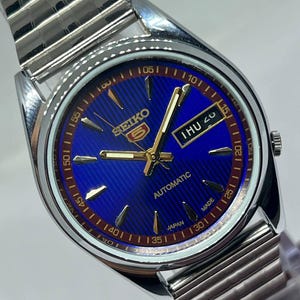 Op de afbeelding: Een zilverkleurig Seiko 5 automatisch polshorloge met een blauwe wijzerplaat. De wijzerplaat heeft goudkleurige wijzers en markeringen, een dag-datumweergave en de tekst "SEIKO 5 AUTOMATIC JAPAN MADE". De band is van zilverkleurig metaal.