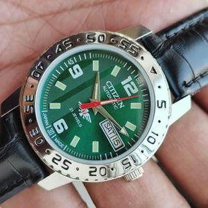 Op de afbeelding: Een Citizen Automatic horloge met een groene wijzerplaat, zilveren lunette en zwarte leren band. De wijzerplaat toont de dag en datum, met lichtgevende markeringen en wijzers. Het horloge is gemarkeerd met "Japan Made".