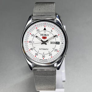 Pode incluir: Um relógio Seiko prateado com mostrador branco e pulseira de malha. O mostrador apresenta algarismos pretos, detalhes vermelhos e o dia da semana. O relógio é marcado como "AUTOMATIC" e "JAPAN MADE."