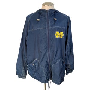 May include: Navy blue hooded jacket with a zipper closure, two front pockets, and white stitching details. The jacket features a yellow and blue "MICHIGAN" logo on the chest. The jacket is made of a lightweight material.