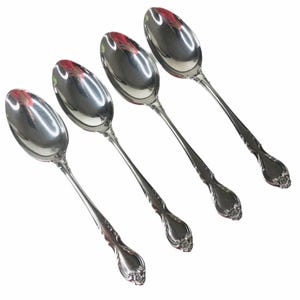 May include: A set of four polished silver-tone metal spoons arranged diagonally. Each spoon features a smooth, oval-shaped bowl and a slender handle that terminates in a decorative, embossed floral motif at the base of the handle.