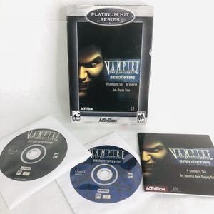 May include: A PC CD-ROM game titled Vampire The Masquerade Redemption Platinum Hit Series. The set includes a game box, two discs in white paper sleeves labeled Disc 1 and Disc 2, and a manual. The cover art features a vampire face.