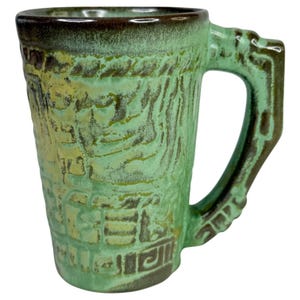 May include: A ceramic mug featuring a textured, relief-style surface with a light green glaze and dark brown accents. The mug has a cylindrical body, a thick handle, and a dark brown rim, showcasing intricate patterns across the exterior surface.