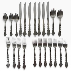May include: A collection of twenty-one stainless steel flatware pieces arranged on a white surface. The set includes seven dinner knives, eight forks, three teaspoons, and two tablespoons, all featuring a matching decorative handle design.
