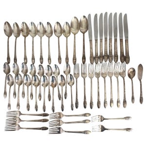 May include: A large collection of silver-toned flatware including various spoons, forks, and knives. The set features ornate, decorative handles with intricate detailing. The pieces are arranged in rows, showcasing a variety of sizes and shapes.
