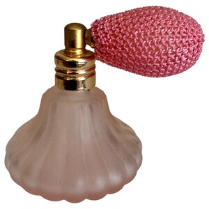 May include: A frosted pink glass perfume bottle with a ribbed, bell-shaped base. The bottle features a gold-toned metal collar and a pink mesh-covered bulb atomizer attached to the side, designed for dispensing fragrance through a pump mechanism.