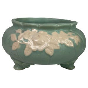 May include: A teal ceramic planter with a floral design. The planter has a rounded shape, three small feet, and a scalloped rim. The floral design is in a lighter cream color and is embossed on the front of the planter.