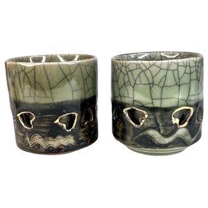 May include: Two ceramic cups with a crackle glaze in shades of green and black. Each cup has heart-shaped cutouts near the base, revealing a gold-colored interior. The cups have a rustic, handcrafted appearance.