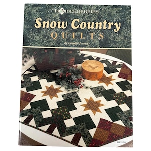 May include: A book titled "Snow Country Quilts" by Lynette Jensen is displayed on a table with a star-patterned quilt. The quilt features brown, green, and cream colors. A small wooden box and a basket with greenery are also on the table.