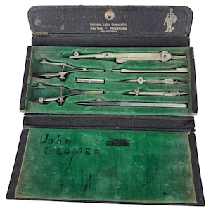 May include: A vintage drafting instrument set in a black case with a green velvet interior. The case features the text Defiance Sales Corporation New York Philadelphia Made in Germany. The interior includes various metal compasses and tools.