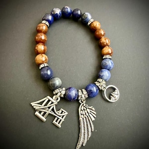 May include: A stretch bracelet featuring alternating blue sodalite and brown wood beads, accented with small silver-toned rhinestone spacers. Three silver-toned metal charms hang from the bracelet: a bridge, a single wing, and a circular mountain icon.
