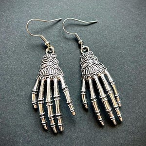 May include: A pair of silver-colored skeleton hand earrings. Each earring features a detailed skeletal hand design, complete with individual finger bones and a textured wrist area. The earrings hang from simple silver hooks.