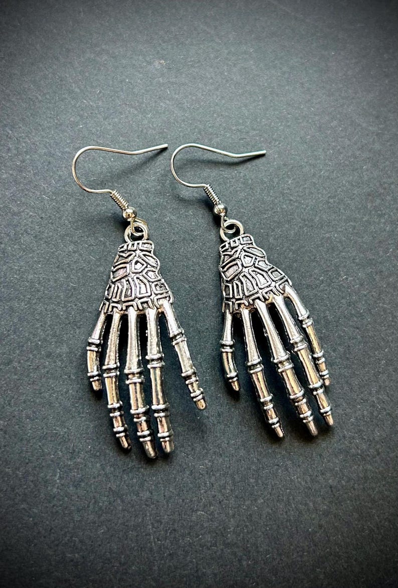May include: A pair of silver-colored skeleton hand earrings. Each earring features a detailed skeletal hand design, complete with individual finger bones and a textured wrist area. The earrings hang from simple silver hooks.