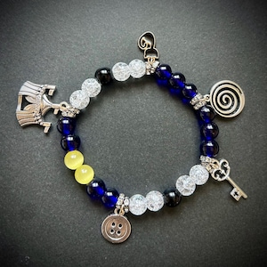 May include: A beaded bracelet with blue, white, black, and yellow beads. The bracelet features silver charms including a key, button, spiral, and a circus-themed emblem. The beads are strung together with silver accents.