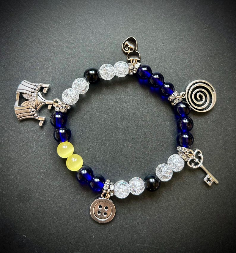 May include: A beaded bracelet with blue, white, black, and yellow beads. The bracelet features silver charms including a key, button, spiral, and a circus-themed emblem. The beads are strung together with silver accents.