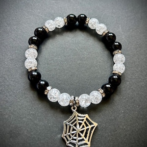 May include: A black and white beaded bracelet with a silver spiderweb charm. The bracelet features alternating black and clear, crackled beads, with small silver accents. The spiderweb charm hangs from the center of the bracelet.